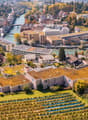 Aerial view of a cityscape with a mix of residential and commercial buildings, surrounded by greenery and a river running through the area.