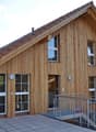 A modern wooden house with a sloped roof, large windows, and a raised ground floor accessed by a staircase.