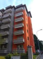 Modern multi-story apartment building with orange and grey facade, multiple balconies, and a gated entrance.