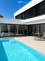 Modern two-story house with large glass sliding doors, a swimming pool, outdoor seating area with lounge chairs and a small table, and a well-maintained garden.