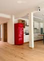 Open-plan kitchen and living area with light wooden flooring, white cabinets, a red refrigerator, a wooden table, and built-in shelving.