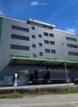 Modern multi-story building with grey facade and green accents, featuring a large truck parked in front and several cars in the parking area.