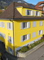 Bright yellow two-story house with white shutters and multiple windows, surrounded by greenery and a neighboring building.