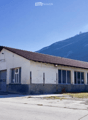 A two-story building with a flat roof and large windows, situated in a rural area with a mountain in the background.