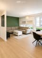 Modern living and dining area with light wood flooring, a beige sectional sofa, a white coffee table, a round wooden dining table with four black chairs, and built-in wooden shelving units.