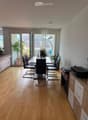 Dining area with a wooden dining table, six chairs, a sofa, a cabinet with electronic devices, and a hanging lamp. Large windows and sliding doors lead to a balcony with plants.