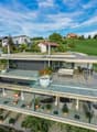 Modern two-story house with a large terrace and glass railings, surrounded by greenery and potted plants. The house features a flat roof and contemporary design.