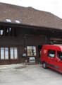 Traditional wooden building with a steeply pitched roof, large windows, and a balcony. A red van is parked in front, and there is a sign on the building.