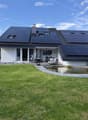 A modern house with solar panels on the roof, surrounded by a well-maintained lawn, a small pond, a patio area with outdoor furniture, and a hot tub.