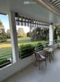 Balcony with white tiled floor, green metal railing, striped awning, small round glass table, and two wicker chairs. Offers a view of a green lawn and trees.