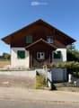 Two-story house with wooden siding, green shutters, and a raised ground floor. Features a garage and small garden area.
