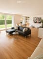 Open-plan living and dining area with wooden flooring, large windows, a black leather sofa, a beige sofa, a coffee table, a dining table with chairs, a kitchen counter, and various decorative items.