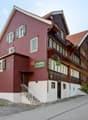 A two-story building with red brick walls and green shutters, featuring a wooden balcony and a sign reading "Mietshaus zur Gögenburg".
