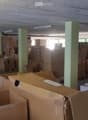 Warehouse space with numerous cardboard boxes stacked on pallets and the floor, green support pillars, and fluorescent lighting.