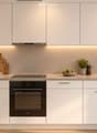 Modern kitchen with white cabinets, stainless steel appliances, and a clean, minimalist design. Features an oven, sink, and countertop with decorative items like potted plants and a bowl of fruit.