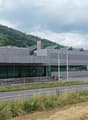 Modern, single-story building with a grey metal facade and large glass windows, situated near a road with a landscaped area and distant hills.