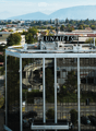 A modern, multi-story building with reflective glass windows and a rooftop sign reading "LUNAJETS." The structure is surrounded by trees, other buildings, and a parking area with several cars.