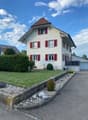 Two-story house with white walls, red shutters, and a well-maintained garden with a gravel pathway.