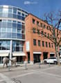 Modern multi-story building with a mix of red brick and glass facade, featuring a rounded glass tower section and several parked cars in front.