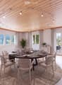 A modern dining room with a round wooden dining table surrounded by six chairs, set with plates, cutlery, and glasses. The room features light wooden flooring, white walls, large windows with curtains, and a sleek cabinet with decorative items. A potted p