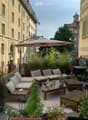 Cozy rooftop terrace with wooden seating, cushions, small table, potted plants, and an umbrella, overlooking a cityscape with historic buildings.
