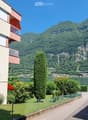 A multi-story residential building with balconies overlooking a scenic lake and mountain backdrop. The area includes greenery, trees, and a paved pathway.