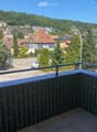 Balcony with a view of a town, featuring tiled flooring and a metal railing, overlooking a street with parked cars and surrounding buildings.