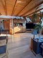Bright living room with wooden floors, exposed wooden beams, large windows, a sofa, a coffee table, a dining table with chairs, a television, and a large potted plant.