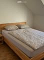 Cozy attic bedroom with wooden bed, nightstand, lamp, and radiator. The bed has a grey duvet and patterned pillows.