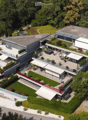 Aerial view of a modern, multi-story residential building with a spacious outdoor area featuring a lawn, patio, and seating arrangements. The building has a sleek design with large windows and a flat roof, surrounded by greenery and neighboring houses.