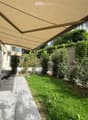 A well-maintained garden area with a covered patio, a covered grill, a small dog, and a variety of plants and shrubs.