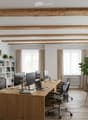 Modern office space with exposed wooden beams, large windows, office desk with multiple monitors, chairs, laptop, printer, bookshelf with books and plants, and a small round glass table with chairs.