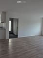 Empty room with wooden flooring, white walls, large windows, and a small kitchen area visible in the background.