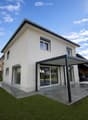 Modern two-story house with white exterior, large windows, and a covered patio area. The house features a well-maintained lawn and is situated in a residential area with neighboring buildings visible in the background.