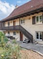 Two-story house with a tiled roof, wooden balcony, and stone facade. Features include multiple windows with shutters, a wooden staircase leading to the entrance, and a small garden area with plants and a bench.