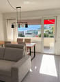 Modern living and dining area with a grey sectional sofa, wooden dining table, chairs, pendant lighting, and large windows offering a scenic view.