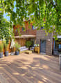Wooden decked outdoor area with a sofa, coffee table, dining table, chairs, and potted plants, surrounded by wooden fencing and trees.