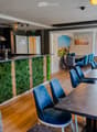 Modern restaurant interior with wooden flooring, blue chairs, tables, a bar counter with a green moss wall, and a seating area with a sofa and bench.