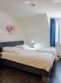 Cozy bedroom with two single beds, a sofa, small side tables with lamps, a large mirror, and a colorful painting on the wall. The room features dark wooden flooring and large windows with blue curtains.
