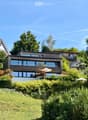Modern two-story house with large windows and wooden exterior, surrounded by lush greenery and trees.