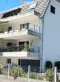 Modern two-story house with white exterior, multiple balconies, and a garage. Features include large windows, a covered balcony area, and solar panels on an adjacent building.