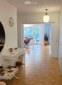 Modern hallway with wooden flooring, white walls, a hanging light fixture, and a round mirror on the wall. A small table with decorative items and a bench are visible, leading to an open area with chairs and a table.