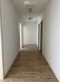 Empty hallway with light wooden flooring and white walls, featuring several doors and ceiling lights.