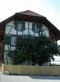 Two-story traditional house with white and brown timber framing, green shutters, and a wooden balcony accessible via stairs.