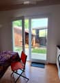 A small, furnished room with a wooden floor, a covered table with a chair, a washing machine, a sink, and a floor lamp. A sliding glass door leads to a backyard.