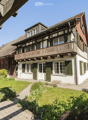 Traditional two-story house with white walls, green shutters, and a wooden balcony. The house features a well-maintained garden with a paved pathway leading to the entrance.