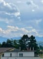 Residential buildings with tiled roofs, surrounded by trees and distant mountains under a partly cloudy sky.