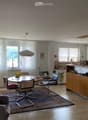 Open-plan living and dining area with a sofa, dining table, chairs, and a modern kitchen featuring white cabinets, a counter, and appliances.