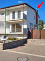 Two-story modern house with white and gray exterior, large windows, balconies, and a wooden gate entrance.