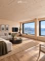 Modern living room with light wooden walls and ceiling, large windows offering a scenic mountain view, a sectional sofa, coffee table, armchair, floor lamp, television, side tables, vases, candles, and picture frames.